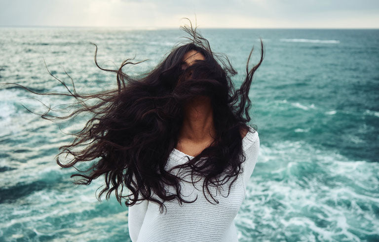 Woman with dark hair flying in the wind with green ocean at the back. Thick shiny lustrous hair with Brahmi Head Massage Oil from Vedic Tiger.